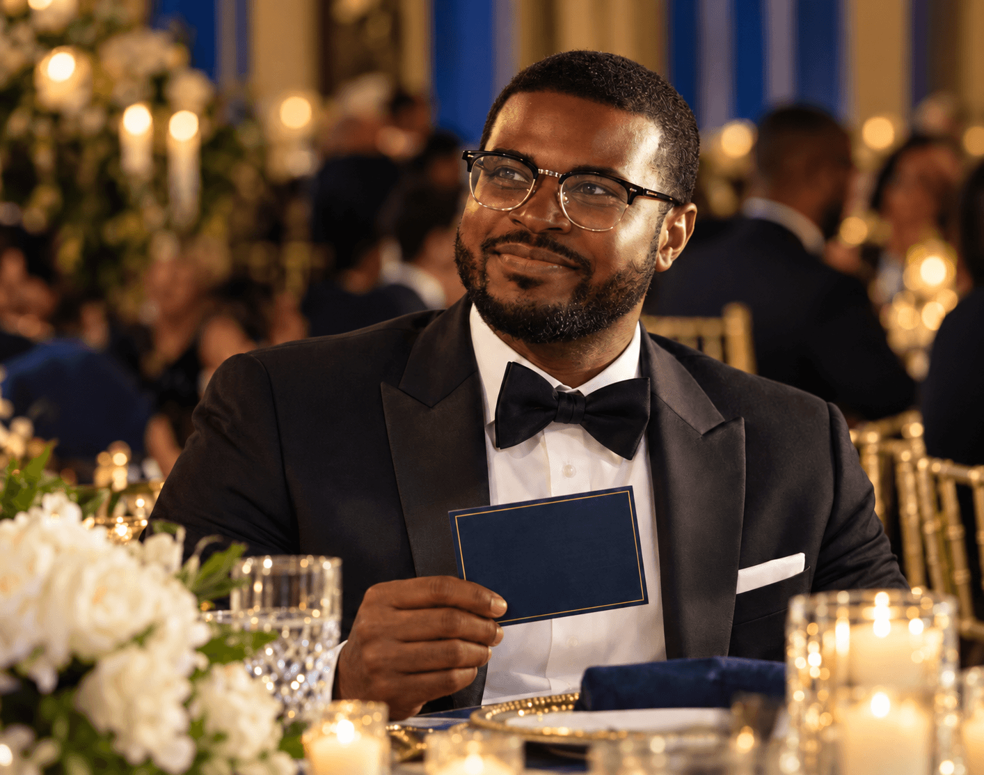 Dennis in a tuxedo at a candlelit gala in Jamaica, holding a blue card with a gold border.